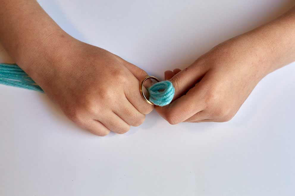 Close-up of hands threading the folded ends of multiple turquoise wool strands through a metal keyring. The wool is being pulled through to form a secure loop.