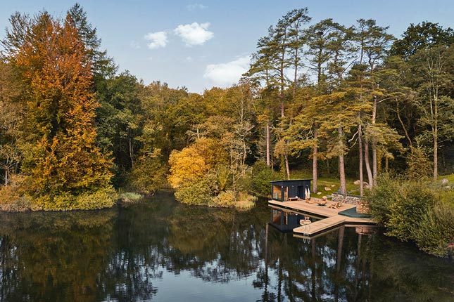 A lake with a wooden jetty with a chill out area and hot tub, surrounded by autumnal trees.