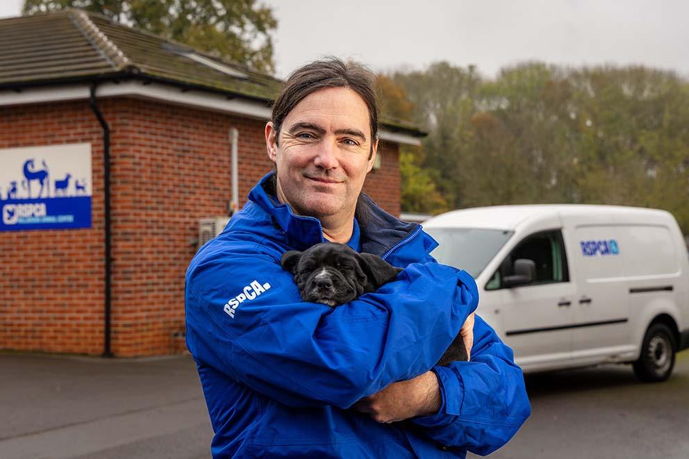 RSPCA Inspector holding small black puppy, he is smilng