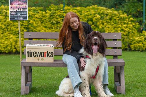 A woman is sat on a bench with a large dog sat between her feet, there is a box and a sign labelled fireworks