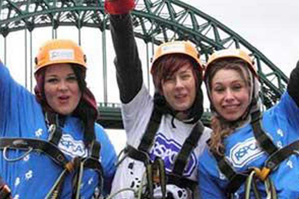 Three women with hands in the air in front of a bridge and wearing abseiling gear.